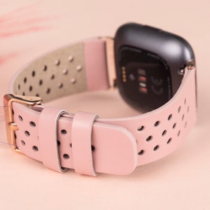 Puede incluir: Una correa de reloj de cuero rosa con un dise&ntilde;o perforado y una hebilla de oro rosa. La correa es compatible con los relojes inteligentes Fitbit Versa.