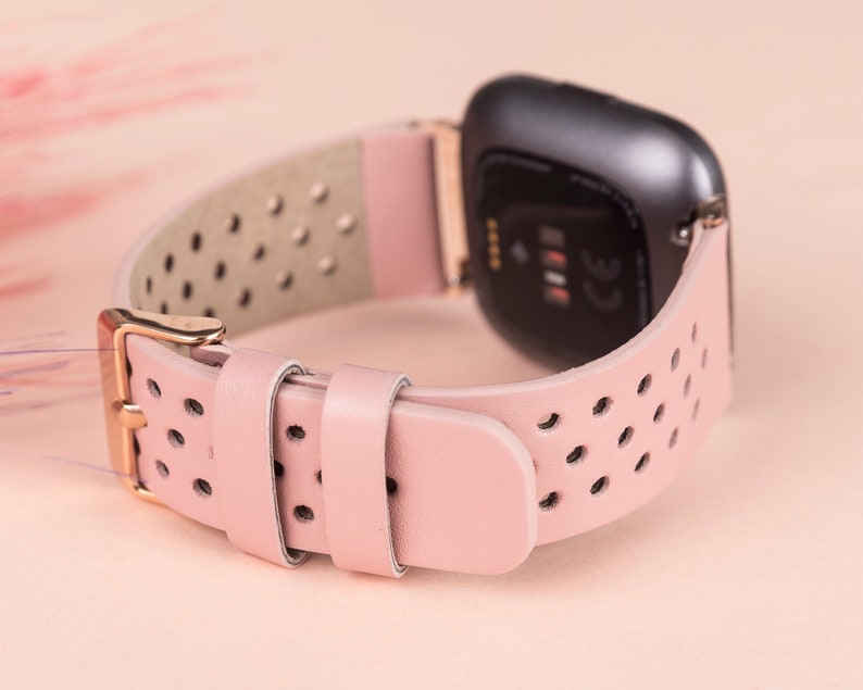 Puede incluir: Una correa de reloj de cuero rosa con un dise&ntilde;o perforado y una hebilla de oro rosa. La correa es compatible con los relojes inteligentes Fitbit Versa.