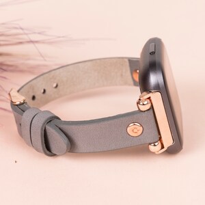 May include: A gray leather watch band with rose gold accents and a black square watch face.