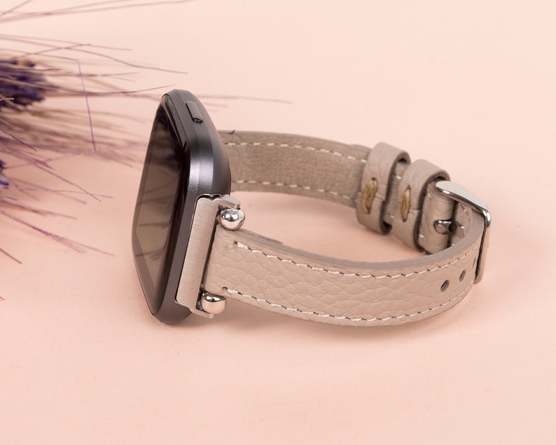 Puede incluir: Una correa de reloj de cuero gris con herrajes plateados y un reloj inteligente negro.