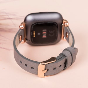 May include: A gray leather watch band with rose gold hardware. The band is designed for a smartwatch with a square face.