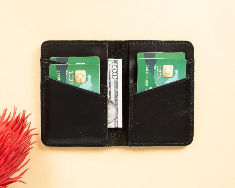 Peut inclure: Portefeuille en cuir noir avec deux emplacements pour cartes et une poche pour l'argent. Le portefeuille est ouvert et contient deux cartes de cr&eacute;dit et un billet d'un dollar.