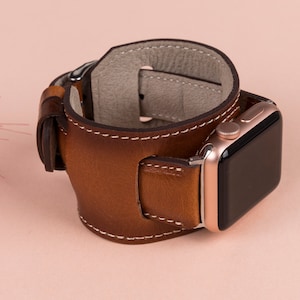 May include: A brown leather watch band with a white stitch detail. The band is designed to wrap around the wrist and has a buckle closure. The band is compatible with Apple Watch.