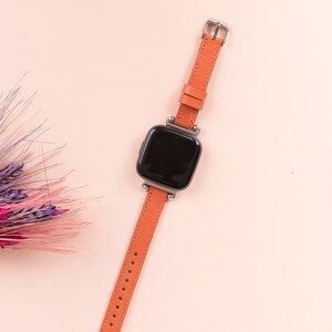 K&ouml;nnte beinhalten: Eine Smartwatch mit schwarzem Zifferblatt und einem leuchtend orangefarbenen Lederarmband. Die Uhr befindet sich auf einem rosa Hintergrund.