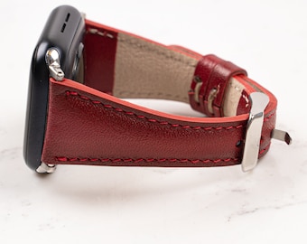 May include: A smartwatch with a dark metallic casing and a deep red leather strap. The strap features visible red stitching along the edges, a silver-toned metal buckle, and a light beige interior lining, showing a textured surface finish.