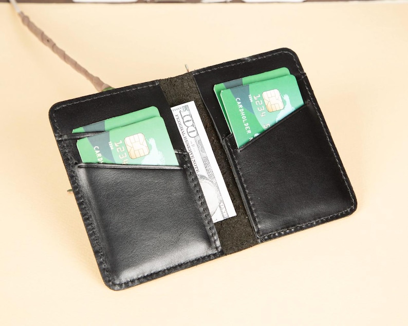 Peut inclure: Un portefeuille en cuir noir avec deux emplacements pour cartes et un porte-billets. Le portefeuille est ouvert et contient deux cartes de cr&eacute;dit et un billet de dollar.