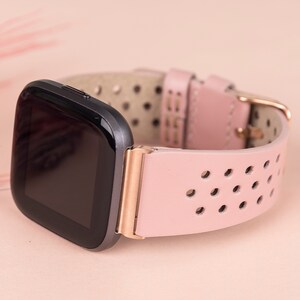 Puede incluir: Una correa de reloj de cuero rosa con hebilla de oro rosa y una esfera de reloj inteligente negra. La correa tiene un dise&ntilde;o perforado.