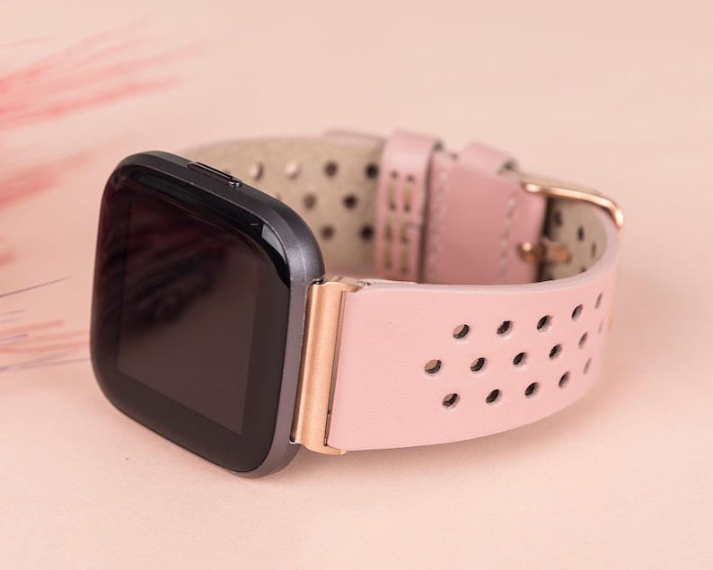 Puede incluir: Una correa de reloj de cuero rosa con hebilla de oro rosa y una esfera de reloj inteligente negra. La correa tiene un dise&ntilde;o perforado.