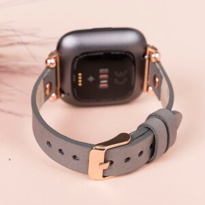 May include: A gray leather watch band with rose gold hardware. The band is designed for a smartwatch with a square face.