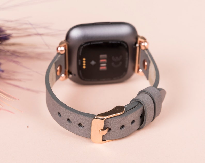 May include: A gray leather watch band with rose gold hardware. The band is designed for a smartwatch with a square face.