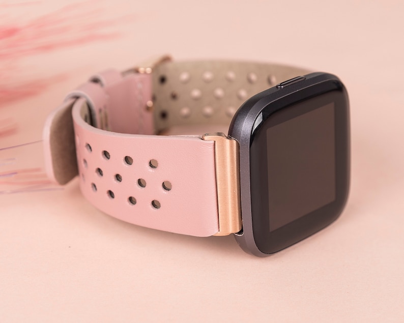Puede incluir: Una correa de reloj de cuero rosa con un acento de oro rosa y una esfera de reloj inteligente negra.