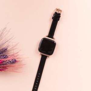 May include: A black smartwatch with a gold-tone buckle and a black leather strap on a pink background.