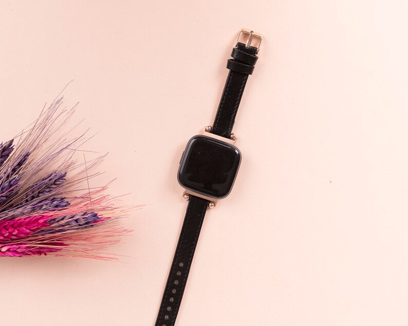 May include: A black smartwatch with a gold-tone buckle and a black leather strap on a pink background.