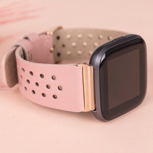 Puede incluir: Una correa de reloj de cuero rosa con un acento de oro rosa y una esfera de reloj inteligente negra.