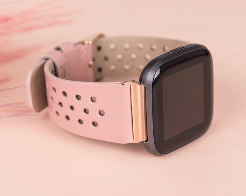 Puede incluir: Una correa de reloj de cuero rosa con un acento de oro rosa y una esfera de reloj inteligente negra.