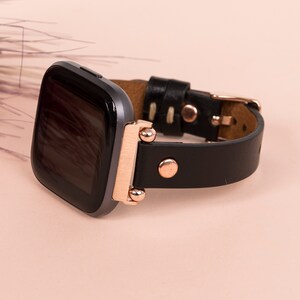 May include: A smartwatch with a dark gray square screen and a black leather strap. The watch features rose gold metal hardware, including the connector lugs, rivets, and a buckle. The strap is secured with two rose gold rivets on each side.