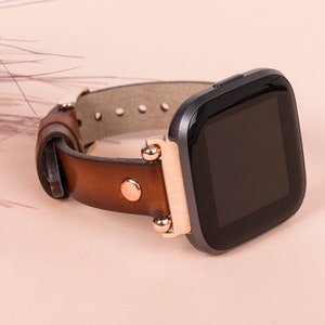 Puede incluir: Una correa de reloj de cuero marr&oacute;n con detalles en oro rosa y un reloj inteligente negro con una esfera cuadrada.