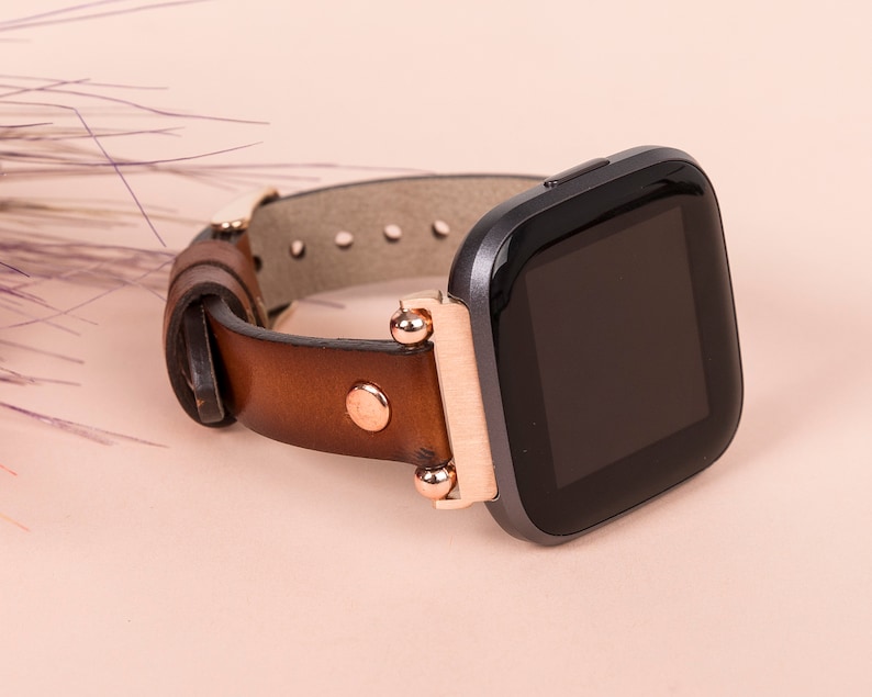 Puede incluir: Una correa de reloj de cuero marr&oacute;n con detalles en oro rosa y un reloj inteligente negro con una esfera cuadrada.