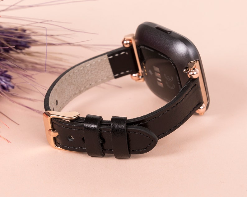 May include: Black leather watch strap with rose gold hardware and a buckle fastening. The strap is compatible with Fitbit Versa smartwatches.