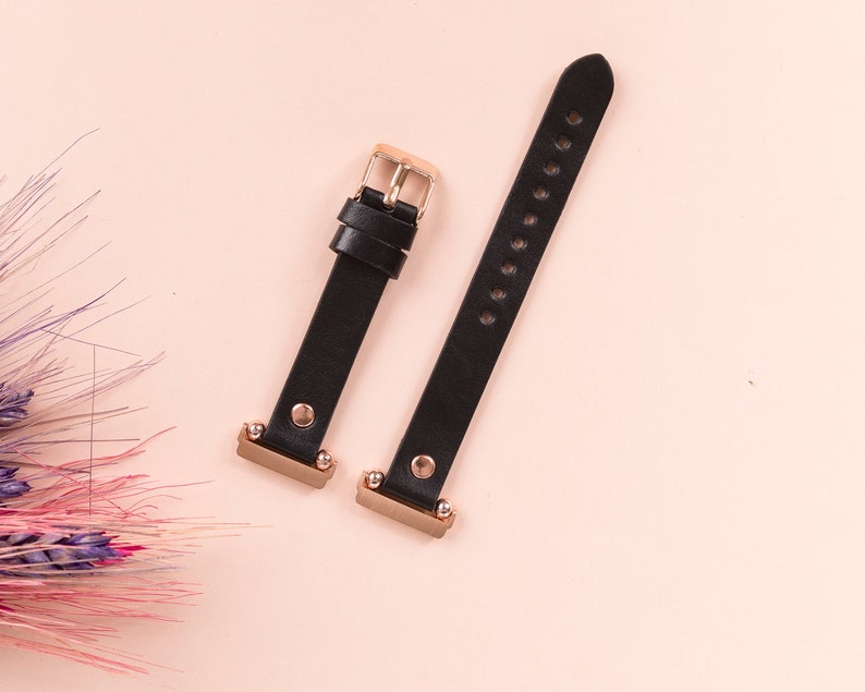May include: A black watch strap consisting of two separate pieces with rose gold hardware. The strap features a rose gold buckle, two matching loops, and rose gold metal connectors at the ends. The leather-like material has a smooth finish.