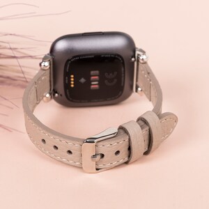 Puede incluir: Un reloj inteligente gris con una correa de cuero gris claro. La esfera del reloj es negra y tiene un peque&ntilde;o punto blanco en el centro.