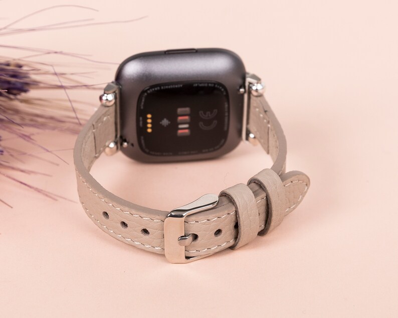 Puede incluir: Un reloj inteligente gris con una correa de cuero gris claro. La esfera del reloj es negra y tiene un peque&ntilde;o punto blanco en el centro.