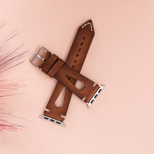 May include: A brown leather watch band with white stitching and a silver buckle. The band is designed for Apple Watches.