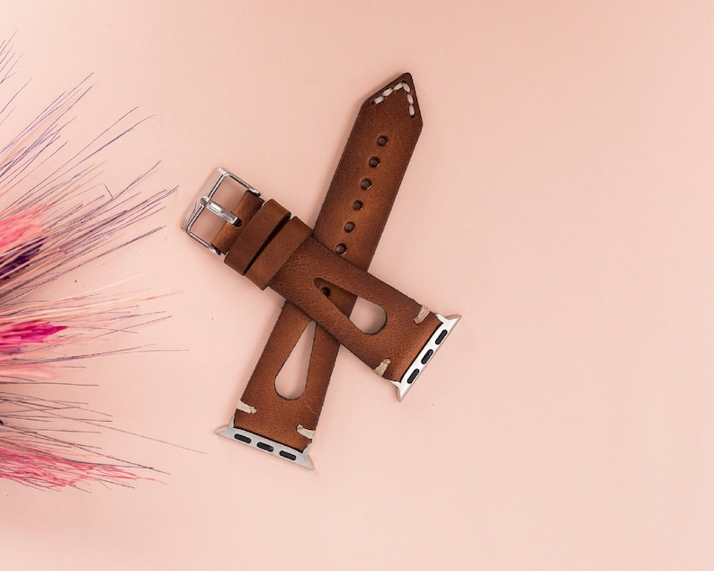 May include: A brown leather watch band with white stitching and a silver buckle. The band is designed for Apple Watches.