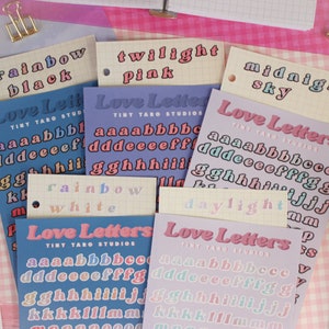 Op de afbeelding: Assortiment van kleurrijke alfabetstickers met de woorden "Love Letters" en "Tiny Taro Studios" op elk vel. De stickers zijn gerangschikt op een roze en witte geruite achtergrond.
