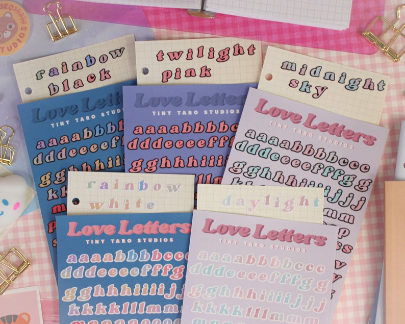 Op de afbeelding: Assortiment van kleurrijke alfabetstickers met de woorden "Love Letters" en "Tiny Taro Studios" op elk vel. De stickers zijn gerangschikt op een roze en witte geruite achtergrond.