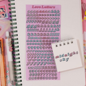 May include: A pink sheet of paper with a set of alphabet stickers in a variety of colors. The stickers are in a retro style font. The text "Love Letters" is at the top of the sheet.  The sheet is on top of a notebook with a grid pattern.