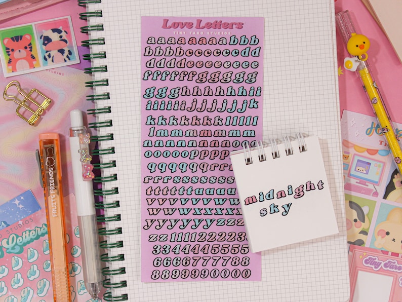 May include: A pink sheet of paper with a set of alphabet stickers in a variety of colors. The stickers are in a retro style font. The text "Love Letters" is at the top of the sheet.  The sheet is on top of a notebook with a grid pattern.