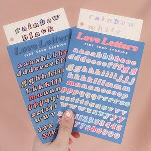 Op de afbeelding: Twee vellen met kleurrijke alfabetstickers met de woorden "Love Letters" en "Tiny Taro Studios" erop gedrukt. De stickers zijn gerangschikt in regenboogkleuren, met de letters "a" tot en met "z" en de cijfers "1" tot en met "9" op elk vel.