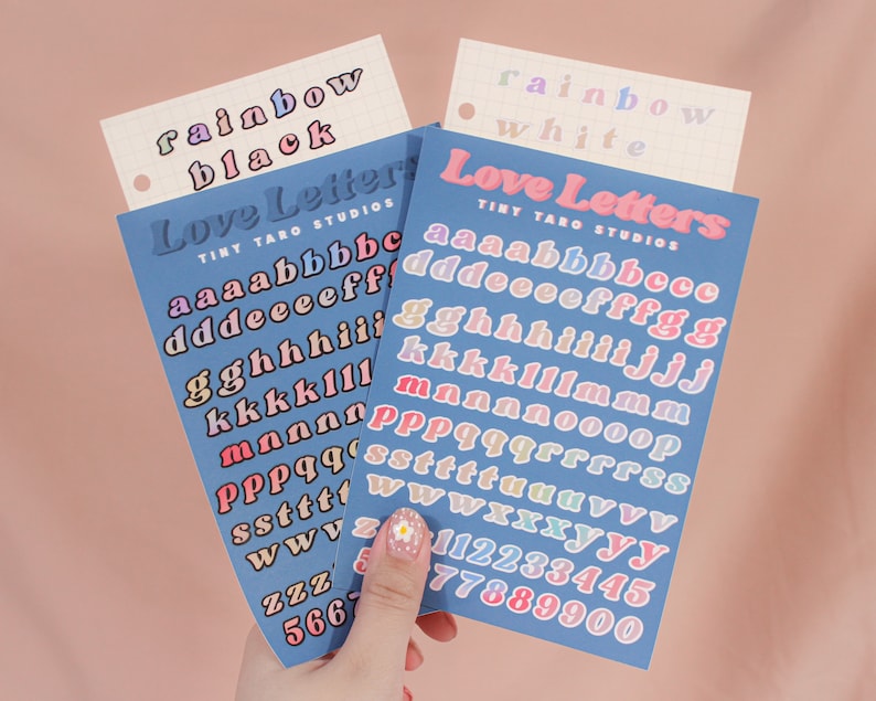 Op de afbeelding: Twee vellen met kleurrijke alfabetstickers met de woorden "Love Letters" en "Tiny Taro Studios" erop gedrukt. De stickers zijn gerangschikt in regenboogkleuren, met de letters "a" tot en met "z" en de cijfers "1" tot en met "9" op elk vel.