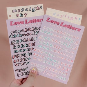Op de afbeelding: Twee vellen stickerpapier met de woorden "Love Letters" en "Tiny Taro Studios" erop gedrukt. De stickers zijn in een pastelkleurenschema en tonen het alfabet in een retro-lettertype. Het eerste vel bevat de letters a-z in roze en blauw, en het tweede vel bevat de letters a-z in roze en blauw, evenals de cijfers 0-9 in roze en blauw.