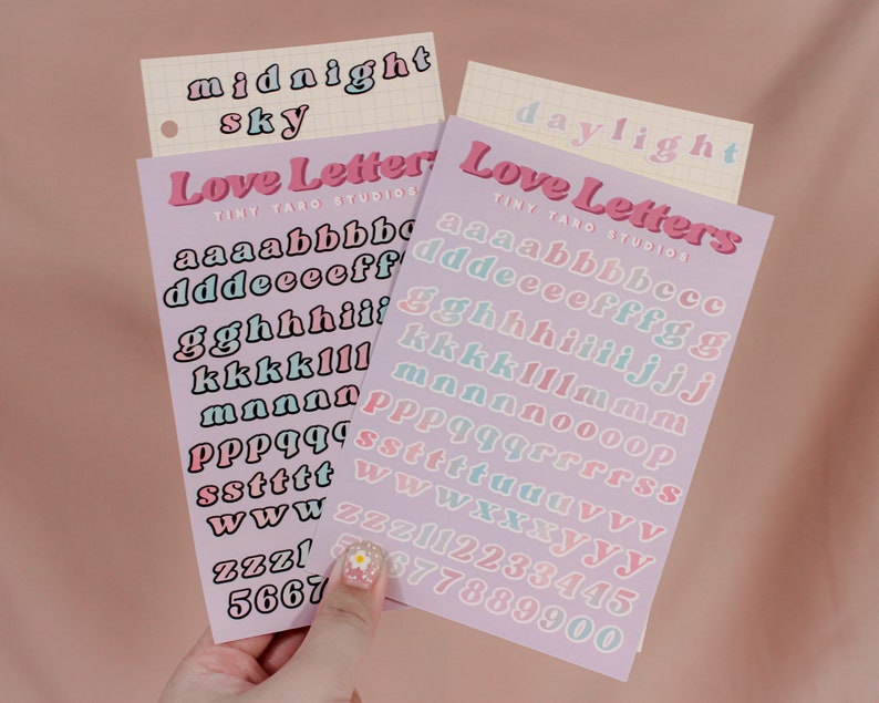 Op de afbeelding: Twee vellen stickerpapier met de woorden "Love Letters" en "Tiny Taro Studios" erop gedrukt. De stickers zijn in een pastelkleurenschema en tonen het alfabet in een retro-lettertype. Het eerste vel bevat de letters a-z in roze en blauw, en het tweede vel bevat de letters a-z in roze en blauw, evenals de cijfers 0-9 in roze en blauw.