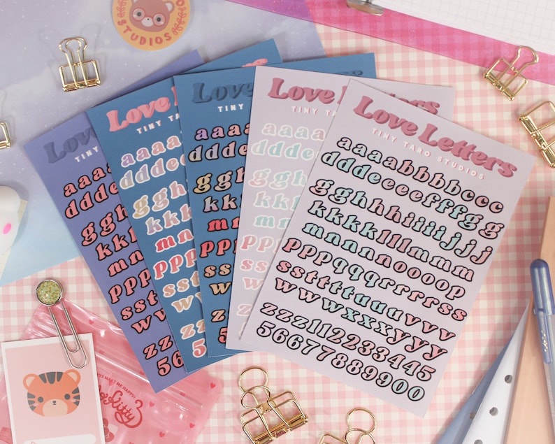 Op de afbeelding: Vier vellen met kleurrijke alfabetische stickers in pastelkleuren. De stickers zijn in een regenboog van kleuren, waaronder roze, blauw, paars en groen. De stickers zijn gerangschikt in rijen, waarbij elke rij een andere letter van het alfabet bevat. De stickers zijn gelabeld met "Love Letters" en "Tiny Taro Studios".