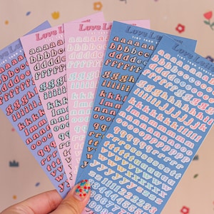 May include: Five sheets of colorful alphabet stickers with a retro font. The stickers are in shades of pink, blue, and yellow. Each sheet has the letters of the alphabet in a different color. The sheets are labeled "Love Letters" and "Tiny Taro Studios".
