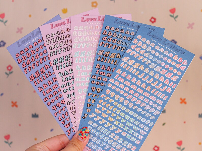 May include: Five sheets of colorful alphabet stickers with a retro font. The stickers are in shades of pink, blue, and yellow. Each sheet has the letters of the alphabet in a different color. The sheets are labeled "Love Letters" and "Tiny Taro Studios".