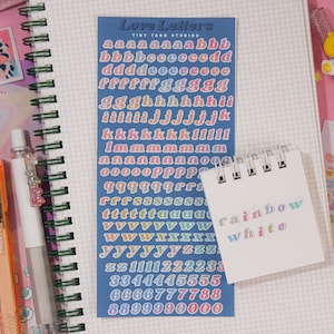 May include: A sheet of colorful alphabet and number stickers on a blue background. The stickers are in a rainbow gradient. The text "Love Letters" and "Tint Taro Studios" is printed on the sheet. A small white notepad with the words "rainbow" and "white" written on it in a rainbow gradient.