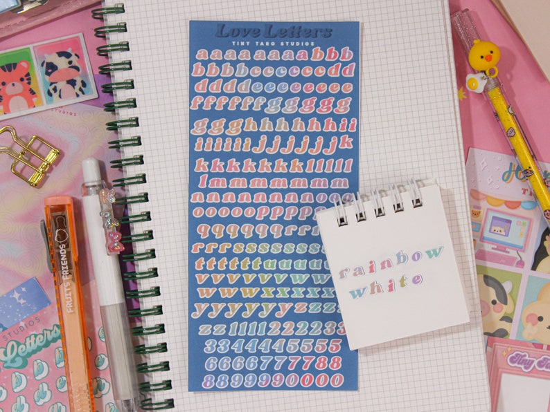 May include: A sheet of colorful alphabet and number stickers on a blue background. The stickers are in a rainbow gradient. The text "Love Letters" and "Tint Taro Studios" is printed on the sheet. A small white notepad with the words "rainbow" and "white" written on it in a rainbow gradient.