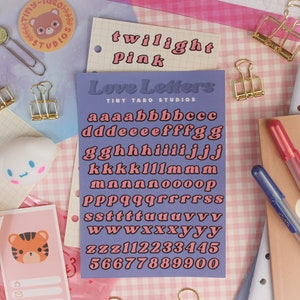 Op de afbeelding: Een paarse kaart met de tekst "Love Letters" en "Tiny Taro Studios" erop gedrukt. De kaart bevat een set roze letters in een retro-lettertype, inclusief het alfabet en de cijfers 1 tot en met 5.
