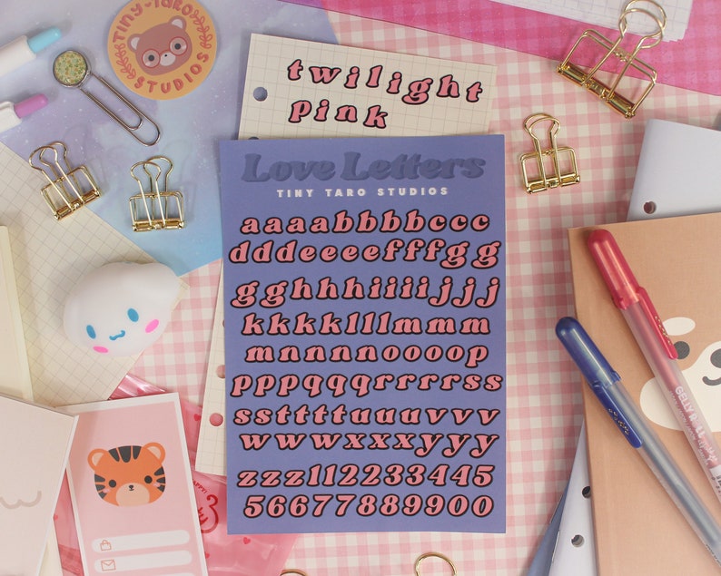 Op de afbeelding: Een paarse kaart met de tekst "Love Letters" en "Tiny Taro Studios" erop gedrukt. De kaart bevat een set roze letters in een retro-lettertype, inclusief het alfabet en de cijfers 1 tot en met 5.