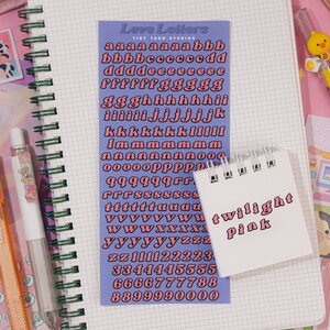 May include: A sheet of pink alphabet stickers with a blue background. The stickers are in a retro style font and include letters, numbers, and punctuation marks. The sheet is labeled "Love Letters" and "Tint Yard Studios". A small white notebook with the words "twilight pink" written on it is in the foreground.