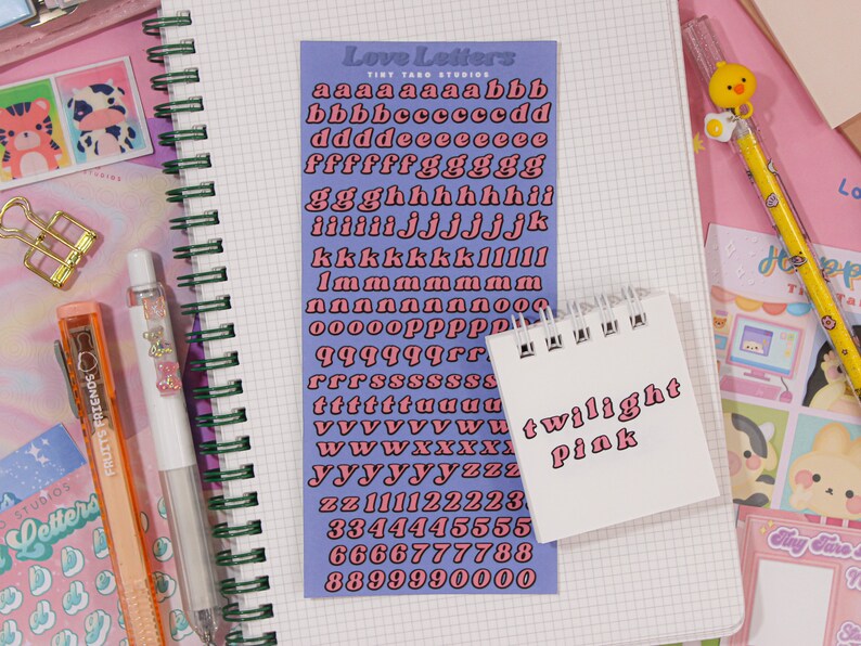 May include: A sheet of pink alphabet stickers with a blue background. The stickers are in a retro style font and include letters, numbers, and punctuation marks. The sheet is labeled "Love Letters" and "Tint Yard Studios". A small white notebook with the words "twilight pink" written on it is in the foreground.