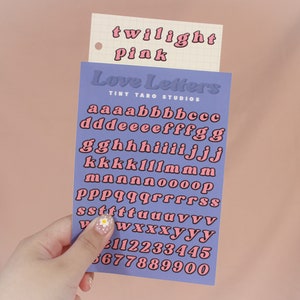 Op de afbeelding: Een vel met roze en zwarte alfabetstickers met de woorden "Love Letters" en "Tiny Taro Studios" erop gedrukt. De letters zijn in een retro-stijl lettertype.