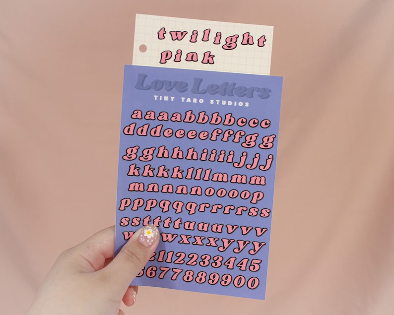 Op de afbeelding: Een vel met roze en zwarte alfabetstickers met de woorden "Love Letters" en "Tiny Taro Studios" erop gedrukt. De letters zijn in een retro-stijl lettertype.