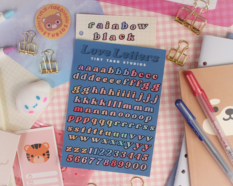 Op de afbeelding: Een blauwe stickervel met de tekst "Love Letters" en "Tiny Taro Studios" in wit. Het vel bevat regenboogkleurige letters, cijfers en symbolen.