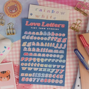 Op de afbeelding: Een vel met kleurrijke alfabet- en cijfer stickers op een blauwe achtergrond. De stickers zijn in een regenboog van kleuren en zeggen "Love Letters" en "Tiny Taro Studios".