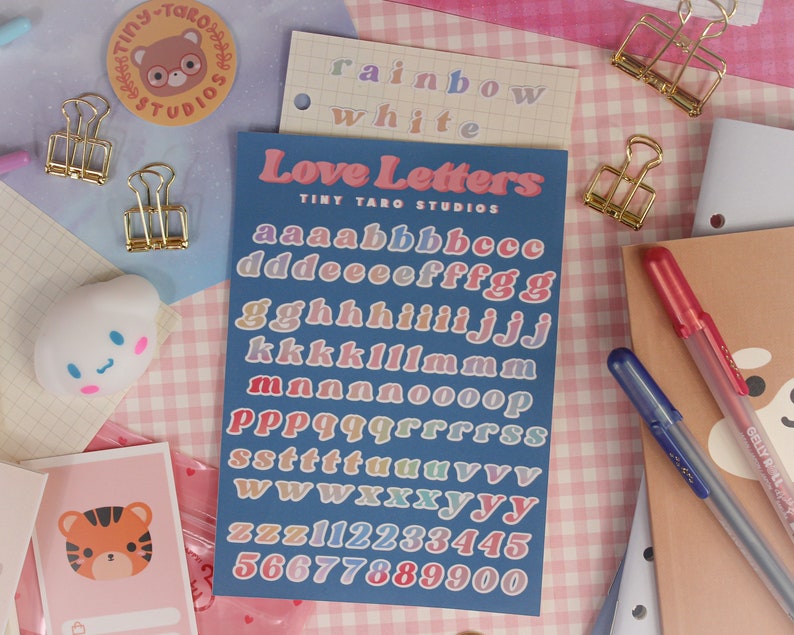 Op de afbeelding: Een vel met kleurrijke alfabet- en cijfer stickers op een blauwe achtergrond. De stickers zijn in een regenboog van kleuren en zeggen "Love Letters" en "Tiny Taro Studios".